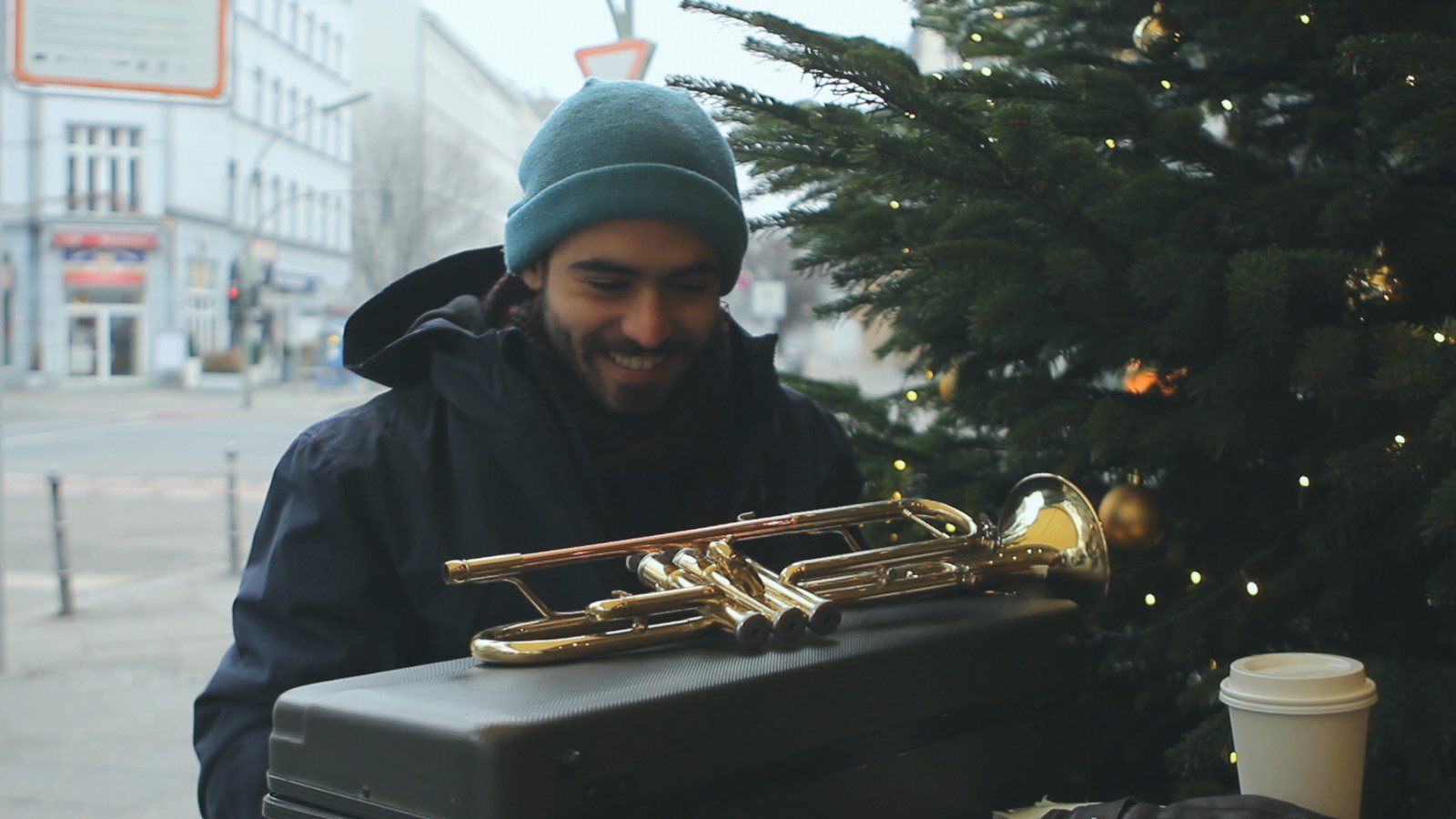 Un homme en terrasse d'un café berlinois en plein hiver, à côté d'un sapin de Noël. L'homme est vêtu d'un bonnet et sourit en découvrant sa trompette après l'avoir sortie de son étui.