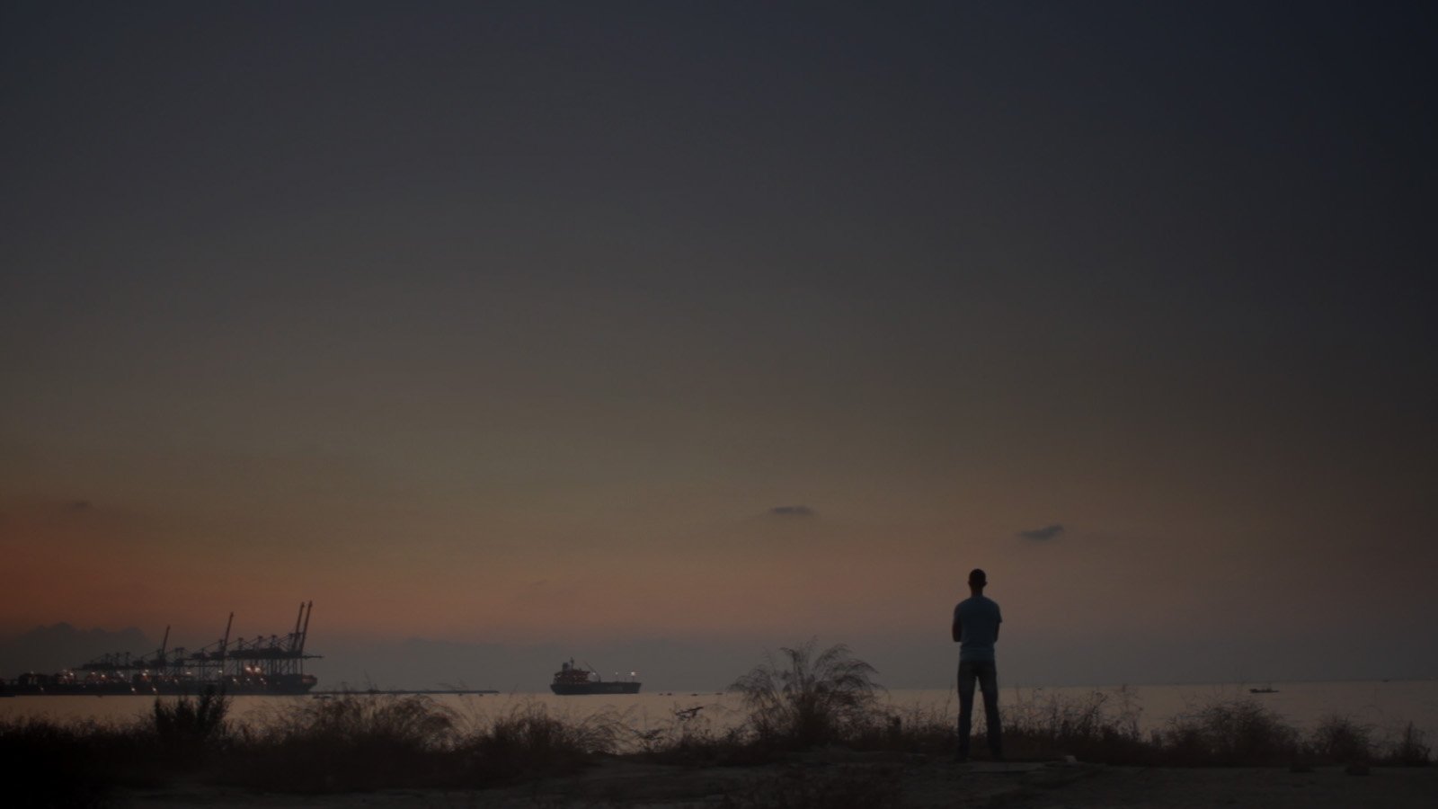 Paysage crépusculaire au bord de la mer, où apparaît la silhouette d'un homme se tenant debout, dos à la caméra et face à la mer.