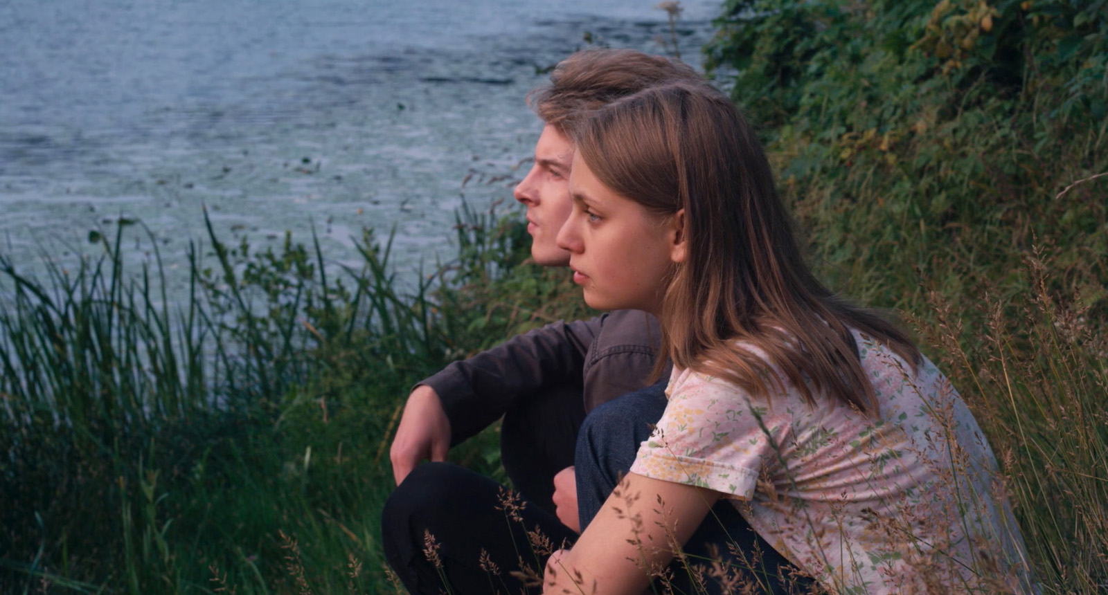 Un jeune couple (une femme et un homme) sont assis dans l'herbe de profil et regardent un lac