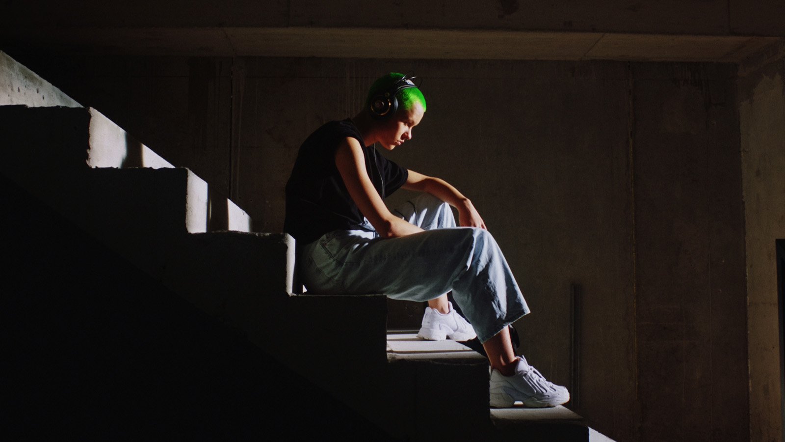 Elina, jeune rappeuse aux cheveux courts et verts, écoute de la musique au casque, assise dans l'ombre d'un escalier